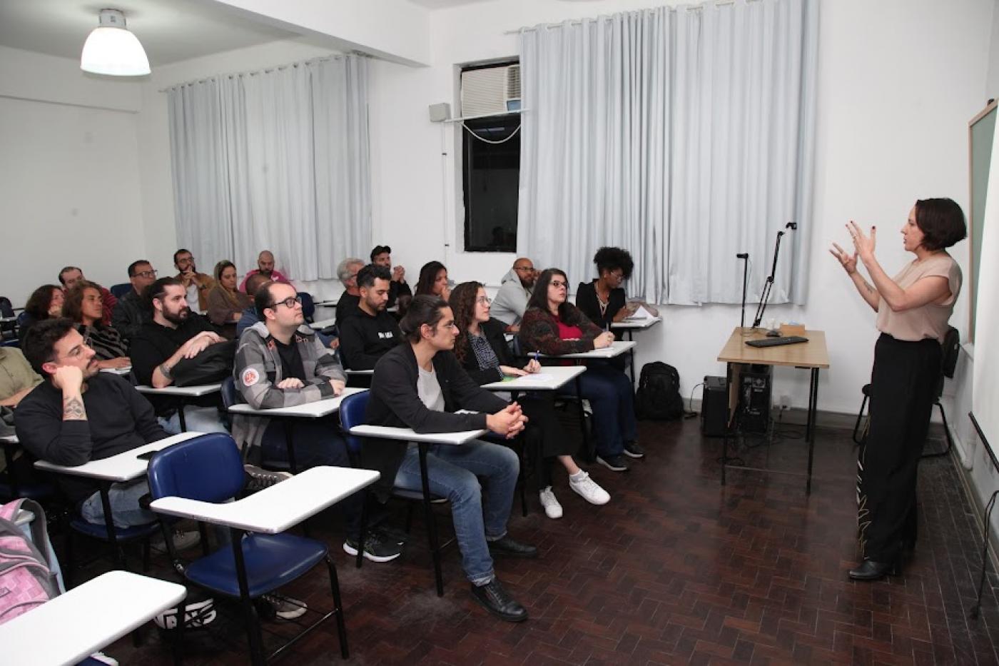mulher está em pé falando a um grupo de pessoas sentadas. #paratodosverem 