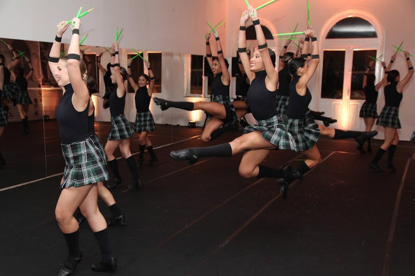 Bailarinas dão salatos no ar. Todas estão seguram um tipo de baqueta em cada mão, ambas levantadas para o alto. #paratodosverem 