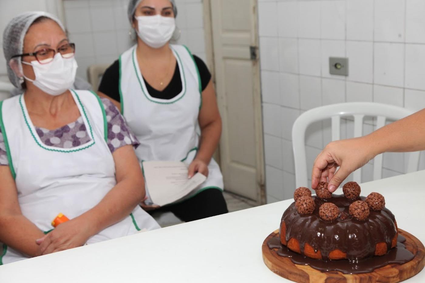 Mão coloca brigadeiro sobre bolo sob olhares de alunas. #paratodosverem