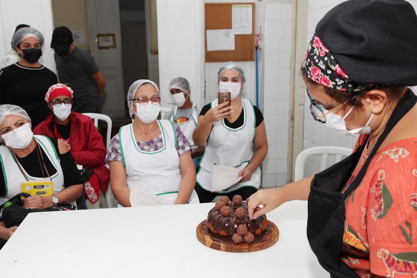 professora decora bolo com brigadeiros. Ao fundo, mulheres assistem à aula. Uma delas fotografa com o celular. #paratodosverem