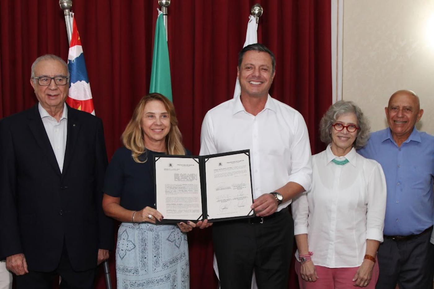 cinco pessoas posam para a foto. Ao centro, o prefeito e a secretária de educação seguram pasta com documento assinado. #paratodosverem 