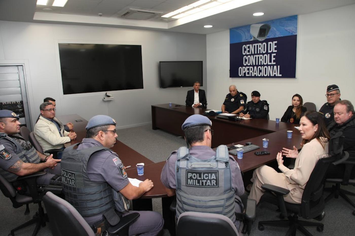 reunião com pessoas sentadas e conversando. Em primeiro plano, dois policiais militares identificados por coletes. #paratodosverem 