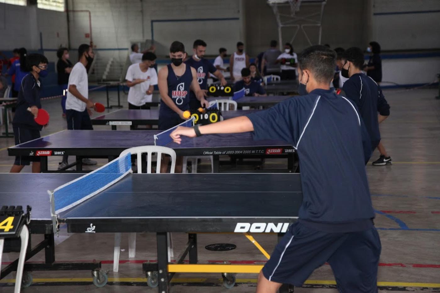 estudantes jogando tenis de mesa em ginásio #paratodosverem
