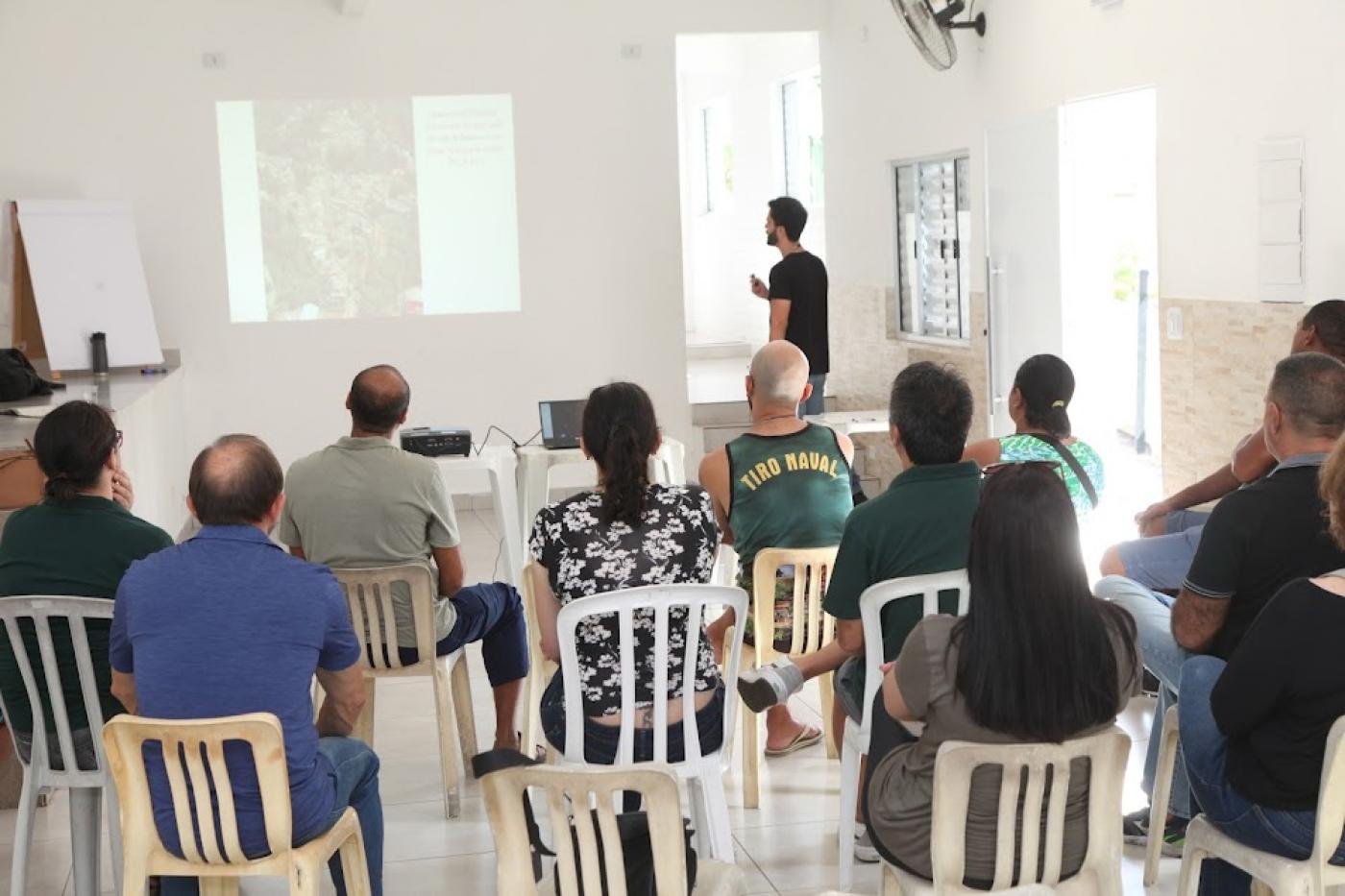 pessoas estão sentadas em sala assistindo a uma apresentação em um telão. #paratodosverem