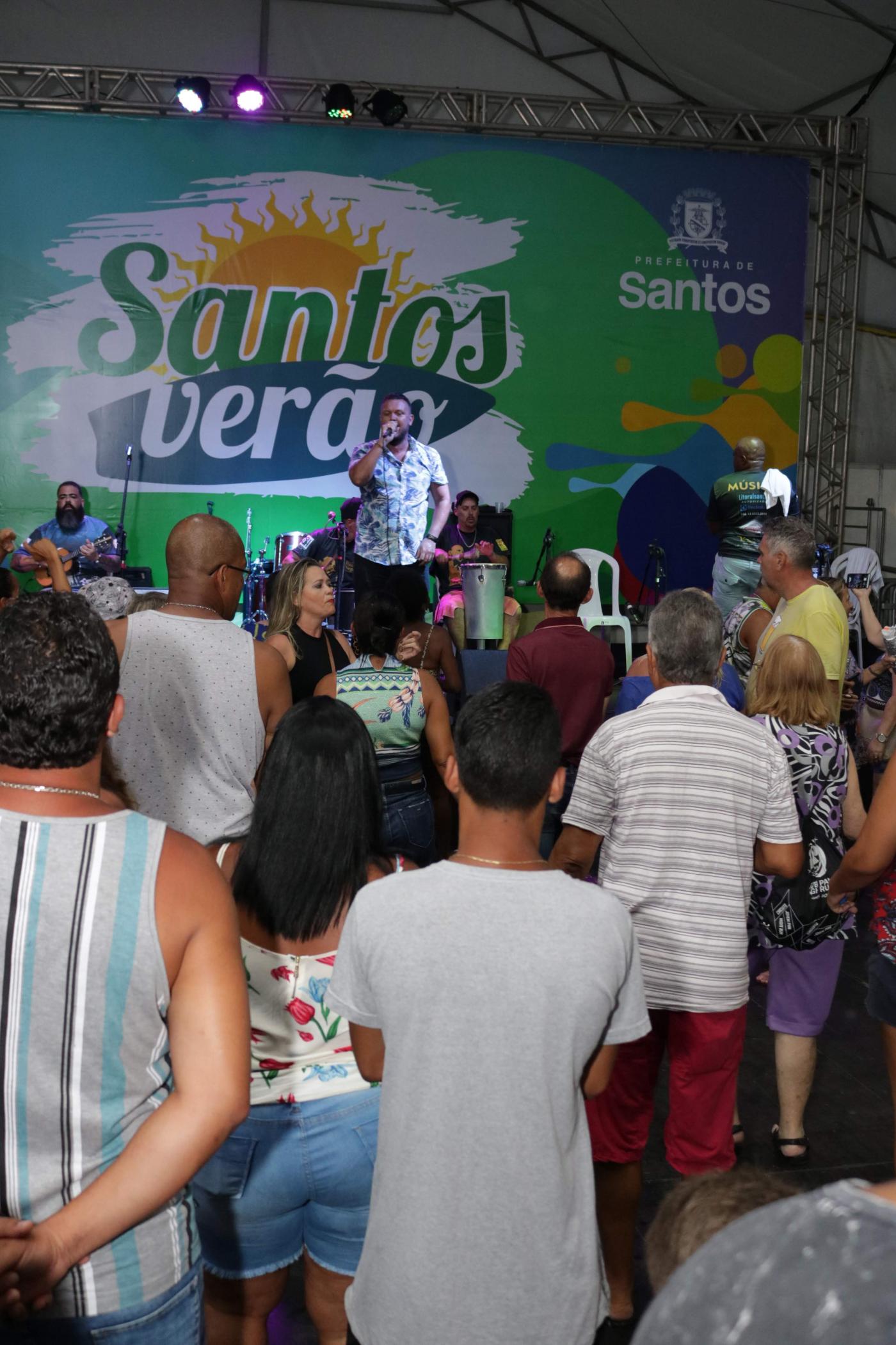 Happy Hour em tenda com cantor ao fundo. Atrás dele há um painel onde se lê Santos Verão. À frente do cantor e de costas para a foto está o  público. #Pracegover