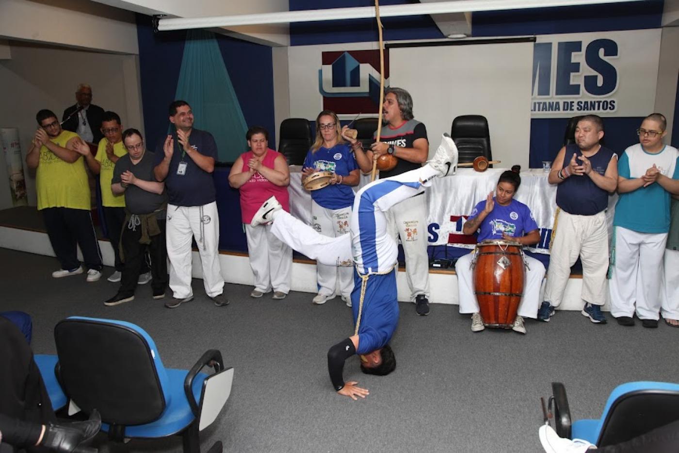 capoeirista estão de cabeça baixo, com as mãos no chão e de pernas para o ar. Atrás, o grupo acompanha com palmas, berimbau e atabaque. #paratodosverem