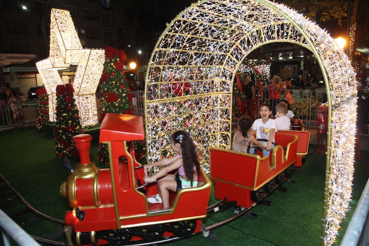 trenzinho circula por trilhos e passa sob túnel de luzes. Crianças dentro do brinquedo sorriem. #paratodosverem 