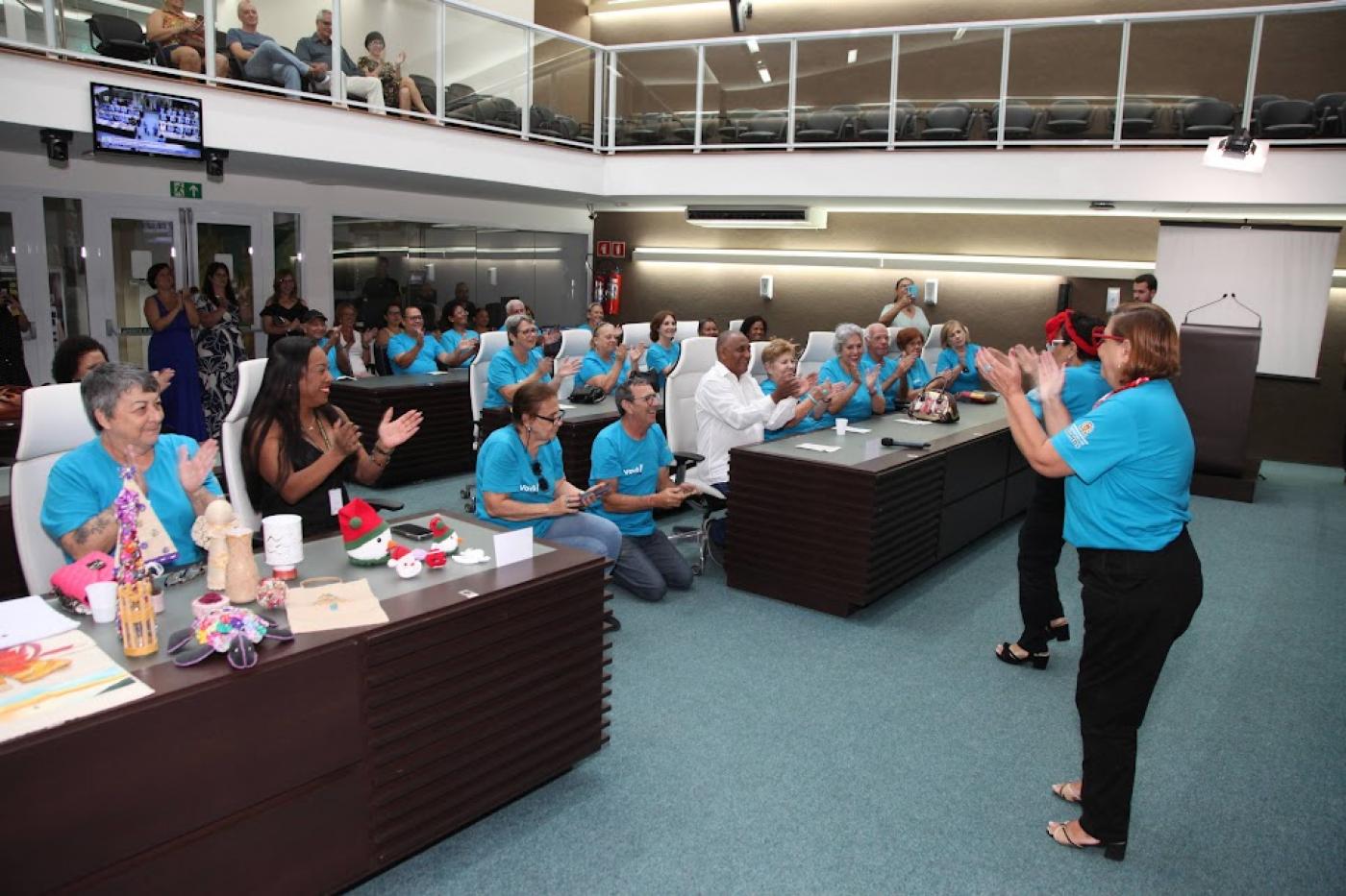 solenidade na câmara com pessoas sentadas e duas mulheres fazendo uma apresentação. Público bate palmas. #paratodosverem 