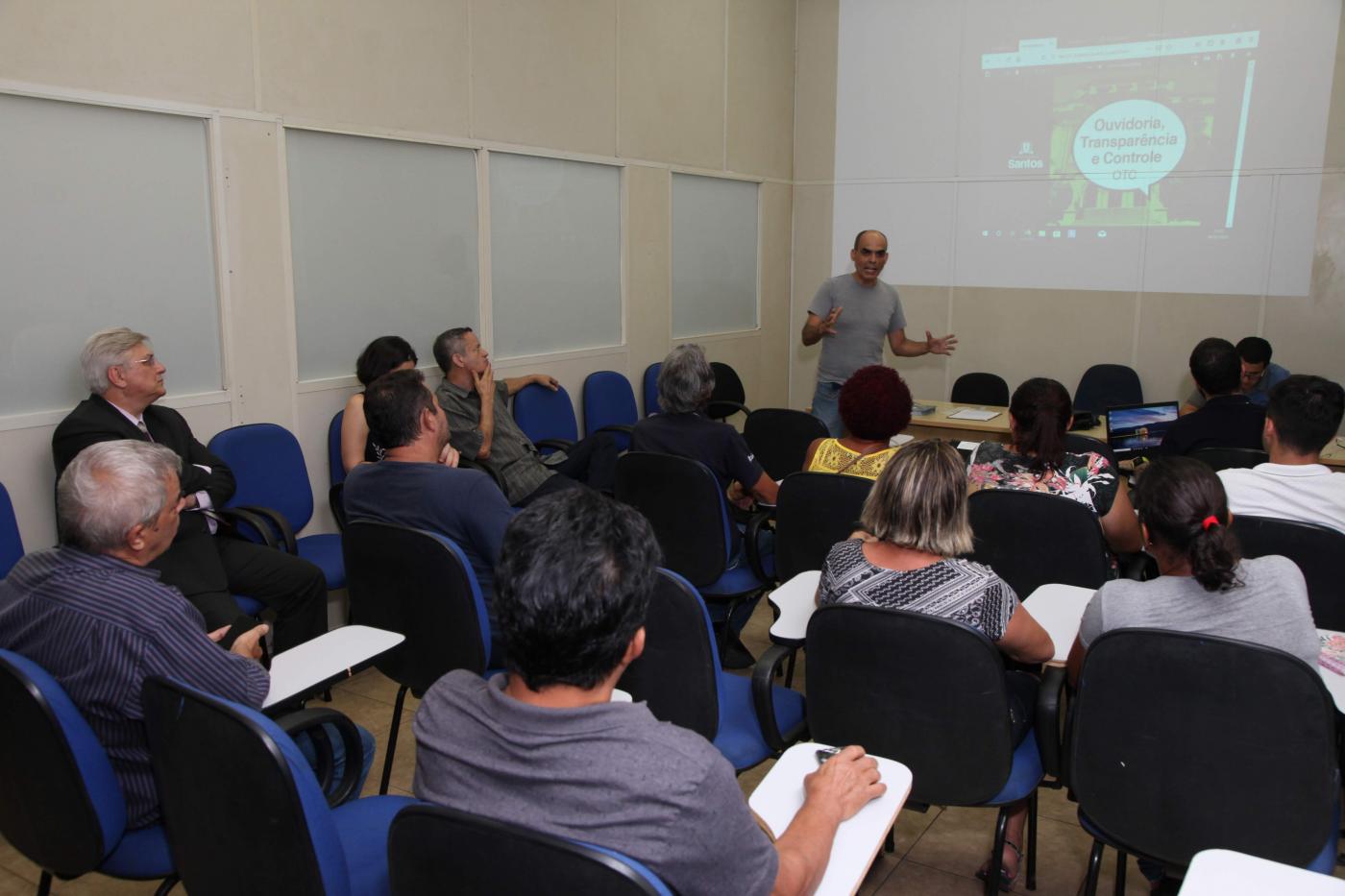 Pessoas estão sentadas em auditório atentas a um homem que fala ao fundo. Há uma imagem projetada na parede do fundo. #Pracegover