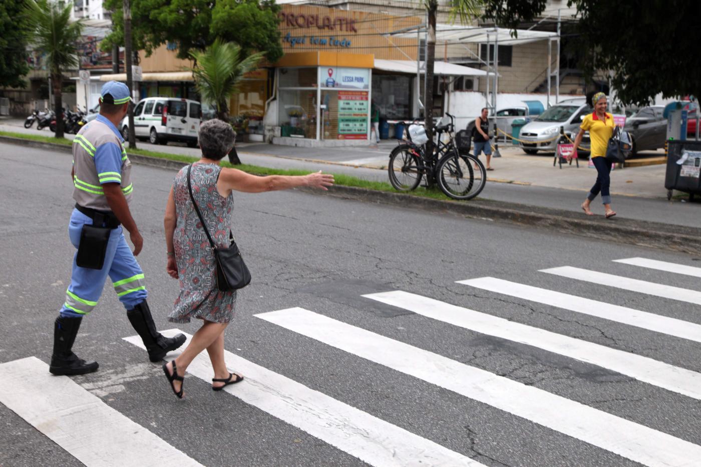 Mulher faz o sinal típico da Faixa Viva. Ela está percorrendo uma faixa de pedestres. Agente da CET está ao lado dela. #Pracegover