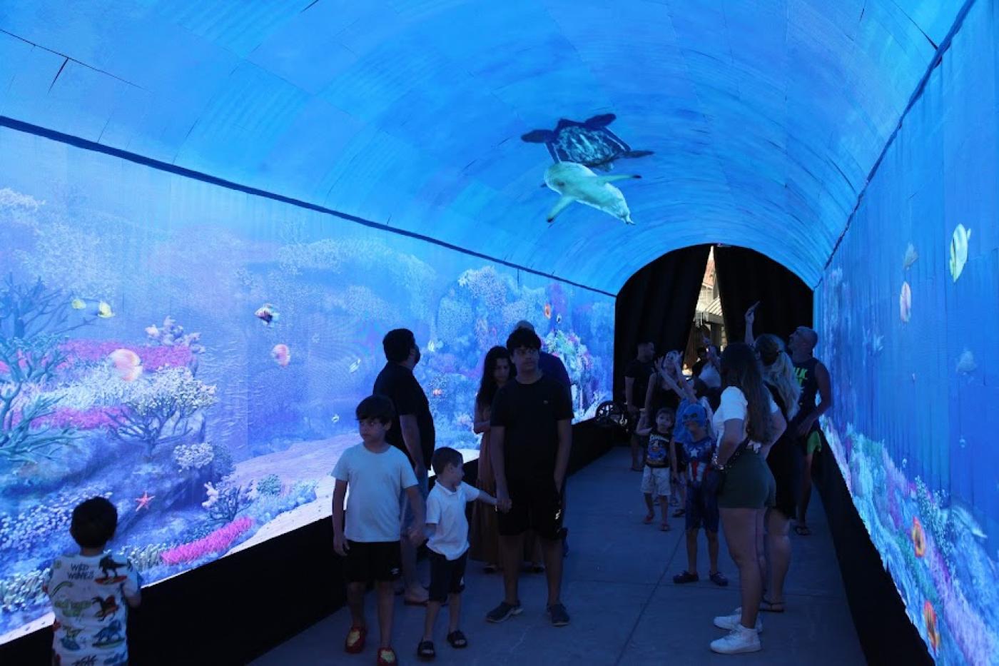 crianças e adultos dentro de túnel apreciando imagens do fundo do mar. #paratodosverem 