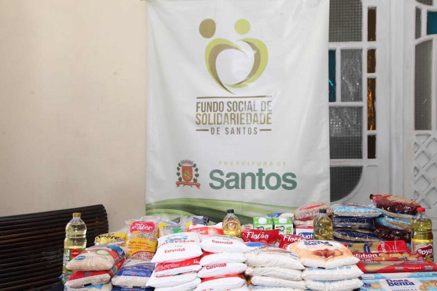 pacotes de alimentos estão sobre uma mesa. Há um banner do fundo social ao fundo. #paratodosverem 