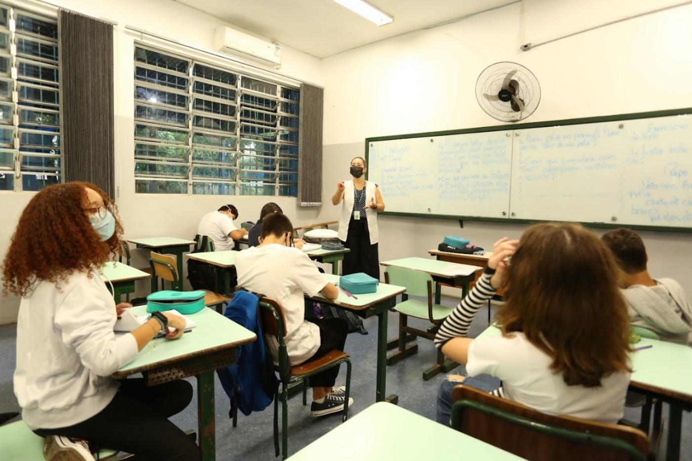 alunos sentados às carteiras estão usando máscara. Ao fundo, a professora à frente do quadro. #paratodosverem