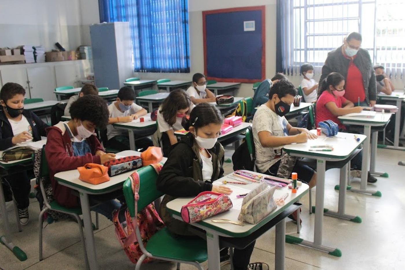 alunos estão sentados em suas respectivas carteiras de classe. Todos usam máscara. A professora está em pé, também com máscara, ao lado de uma aluna sentada. #paratodosverem