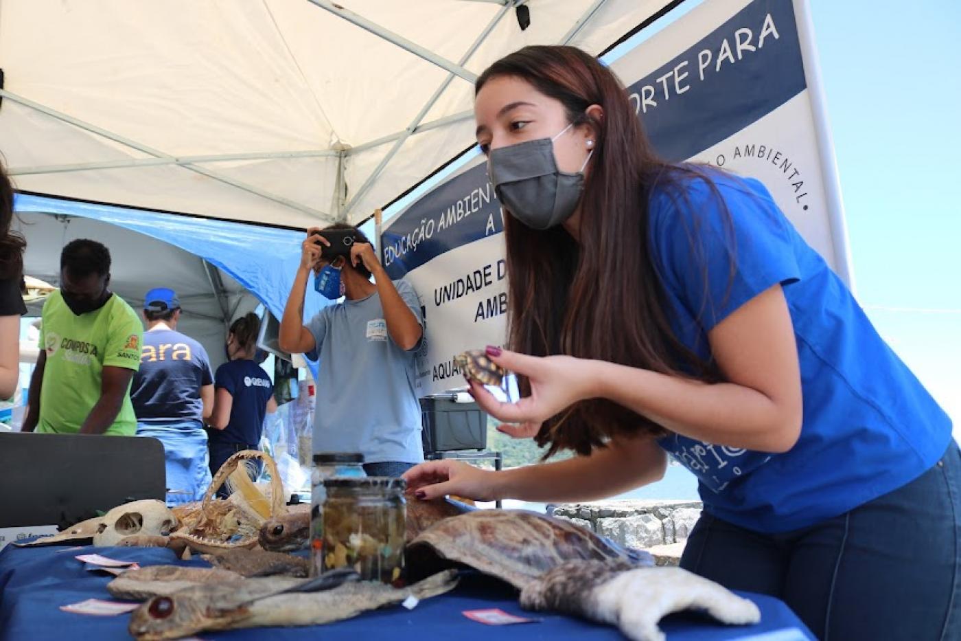 Jovem está atrás de mesa de investigação marinha formada por ossos de peixes, cascas de tartaruga e outros itens. #paratodosverem