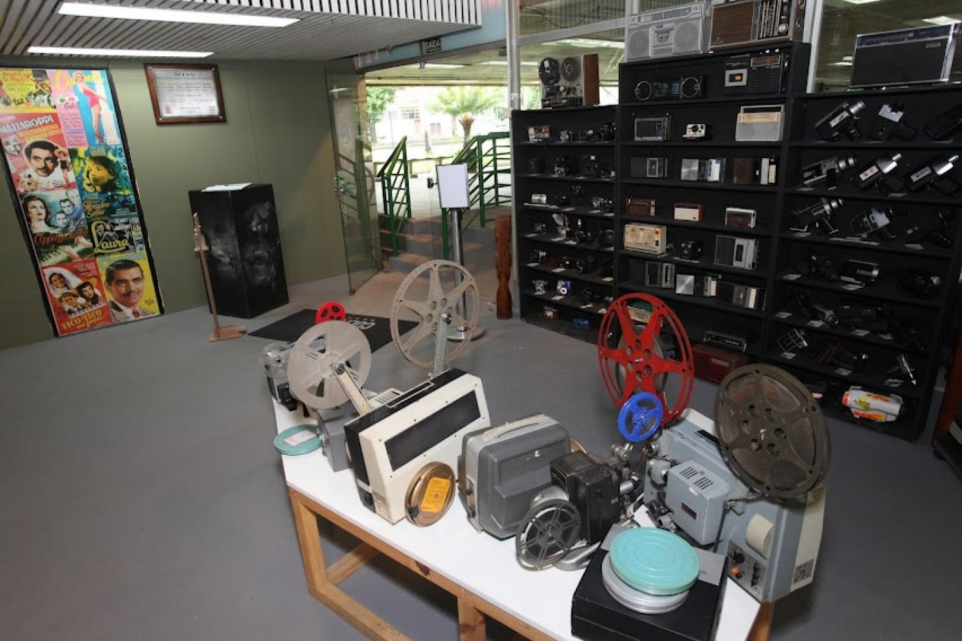 área interna de museu. com mesa e vários objetos antigos, como câmeras e filmadoras sobre uma mesa. Ao fundo, prateleiras com objetos de fotografia. #paratodosverem