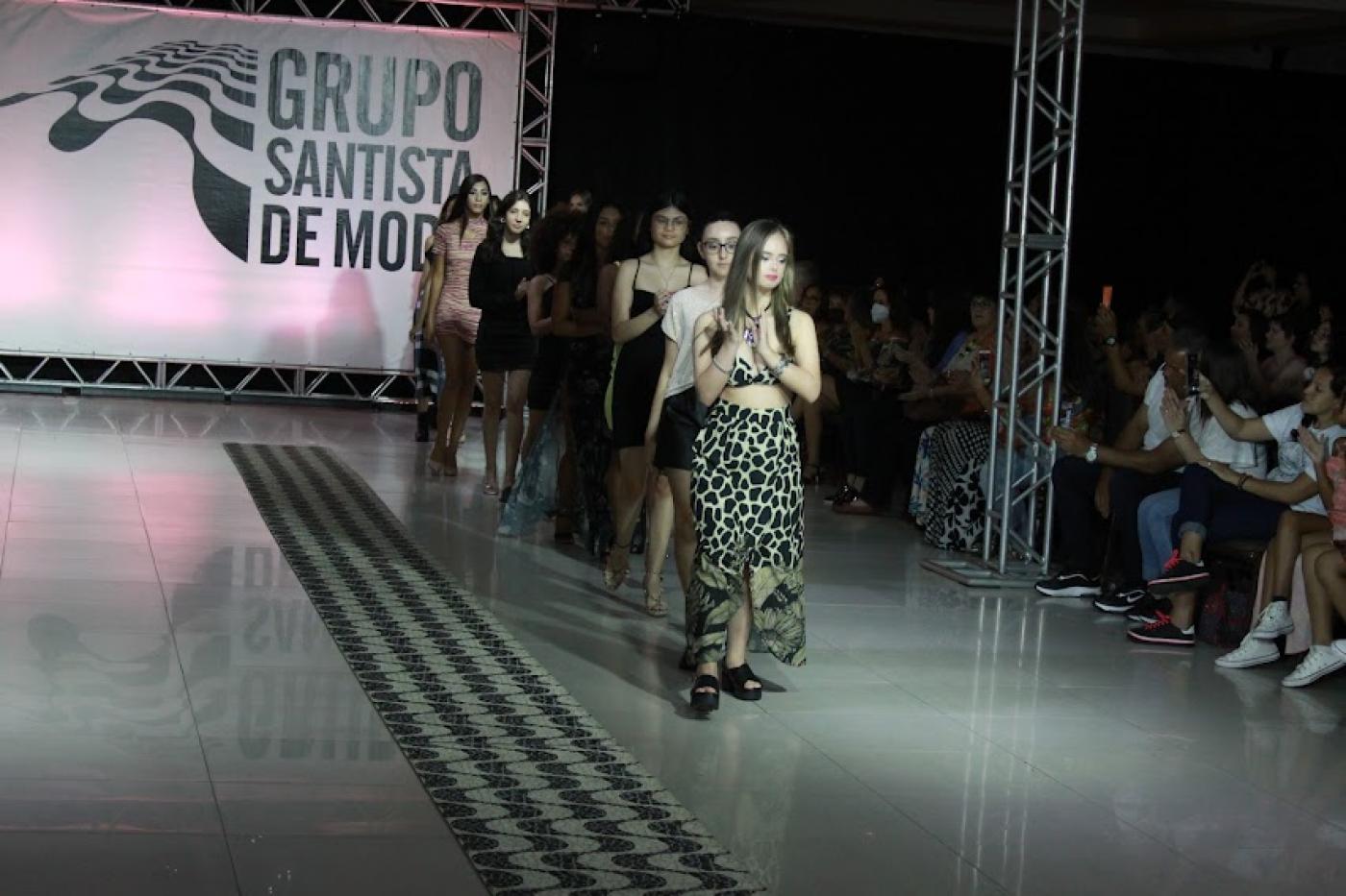 Jovens modelos femininas desfilam na passarela com letreiro ao fundo escrito Grupo Santista de Moda. #pratodosverem