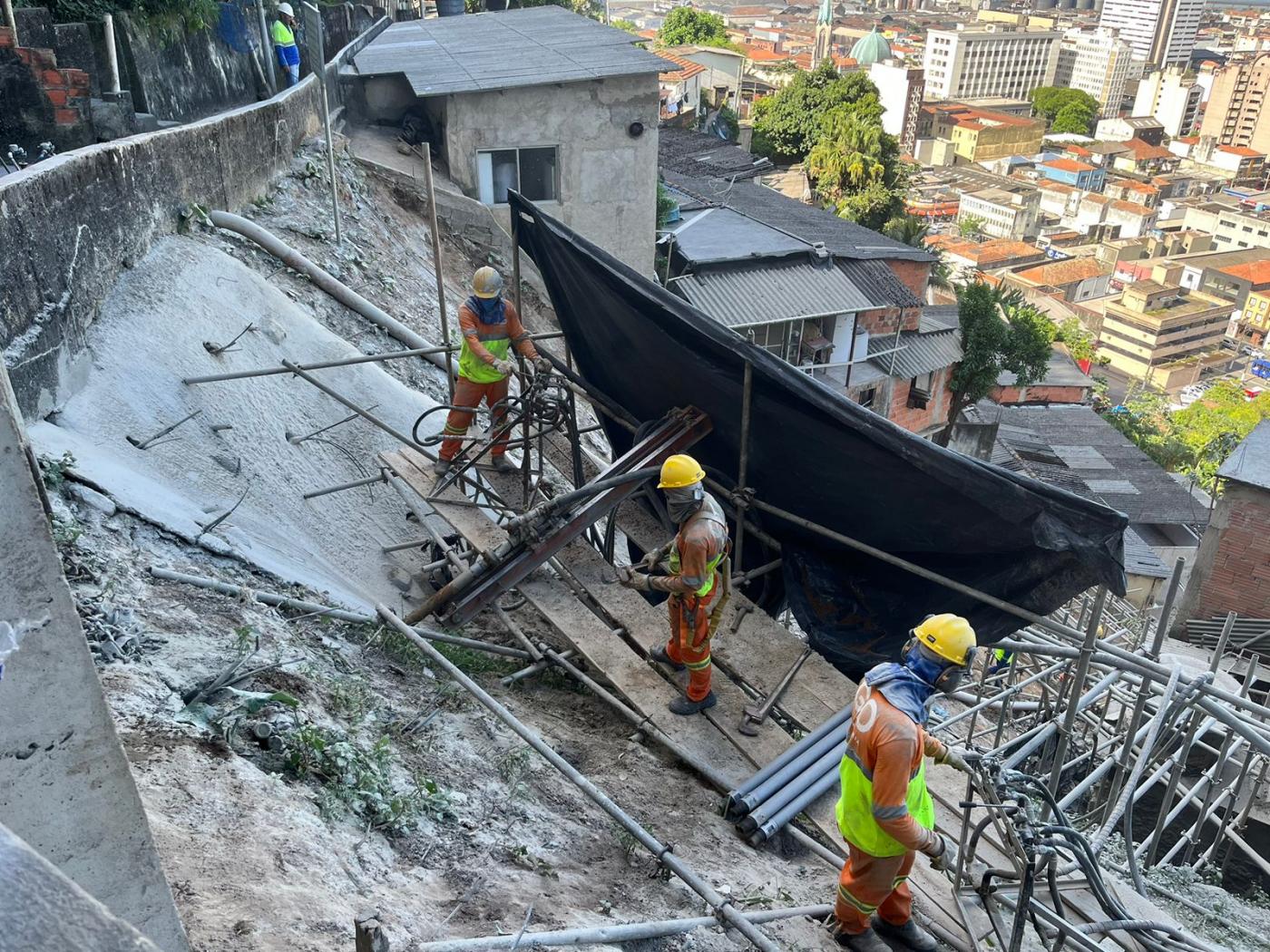 homens estão trabahando em encosta em meio a estruturas de ferro. Há moradias abaixo. #paratodosverem