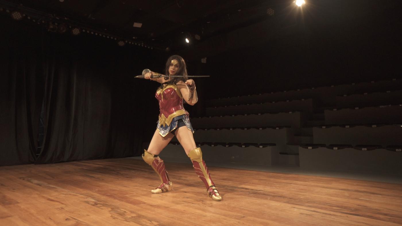 Menina está vestida de mulher-maravilha e faz pose empalco. #paratodosverem