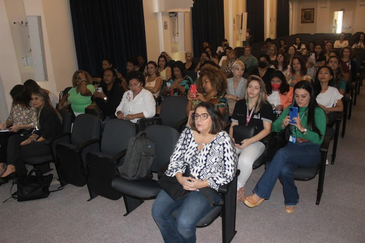 mulheres estão sentadas em auditório. #paratodosverem