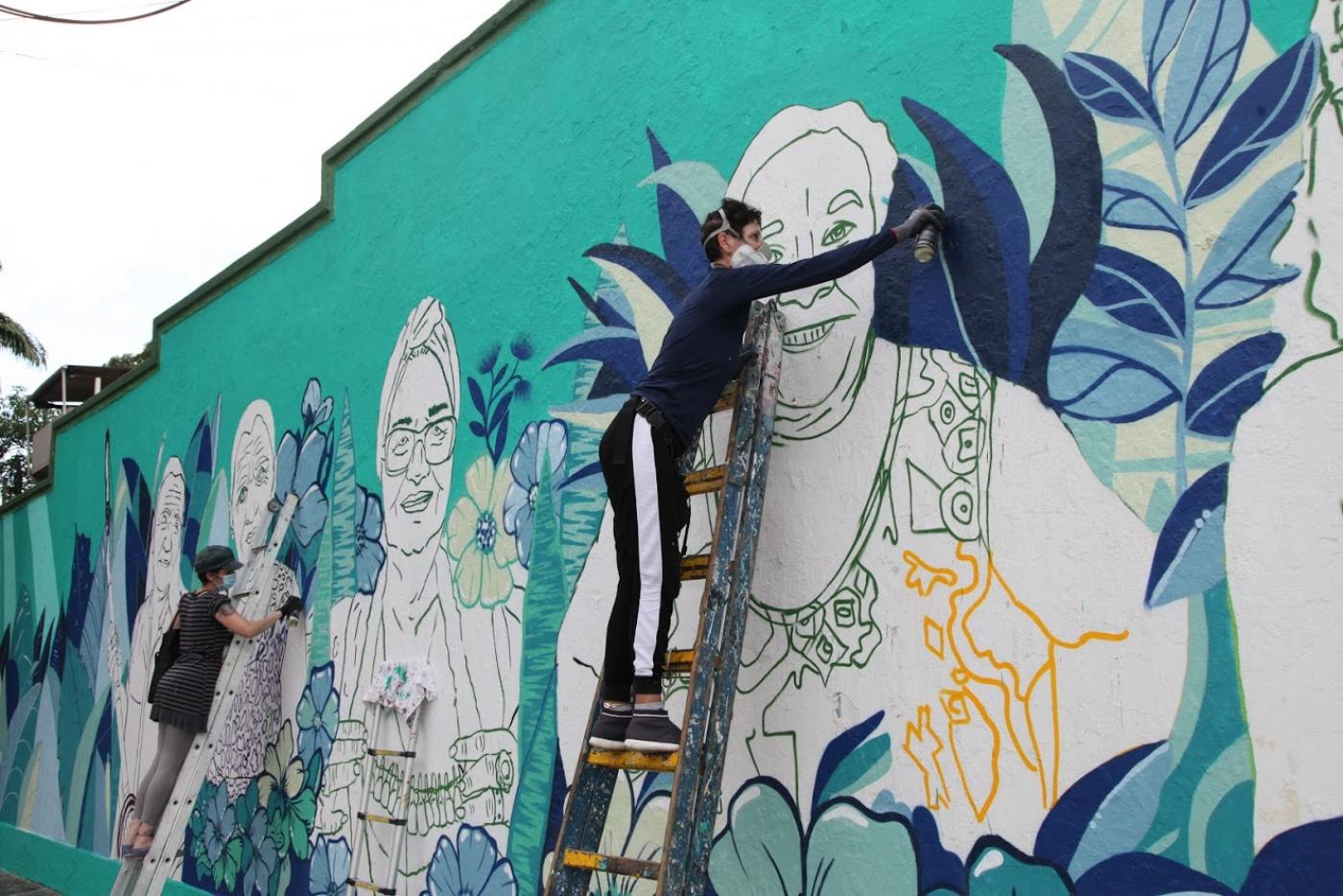 Duas artistas estão no alto de escadas encostadas a um muro. Elas desenham e grafitam imagens de mulheres. #Paratodosverem