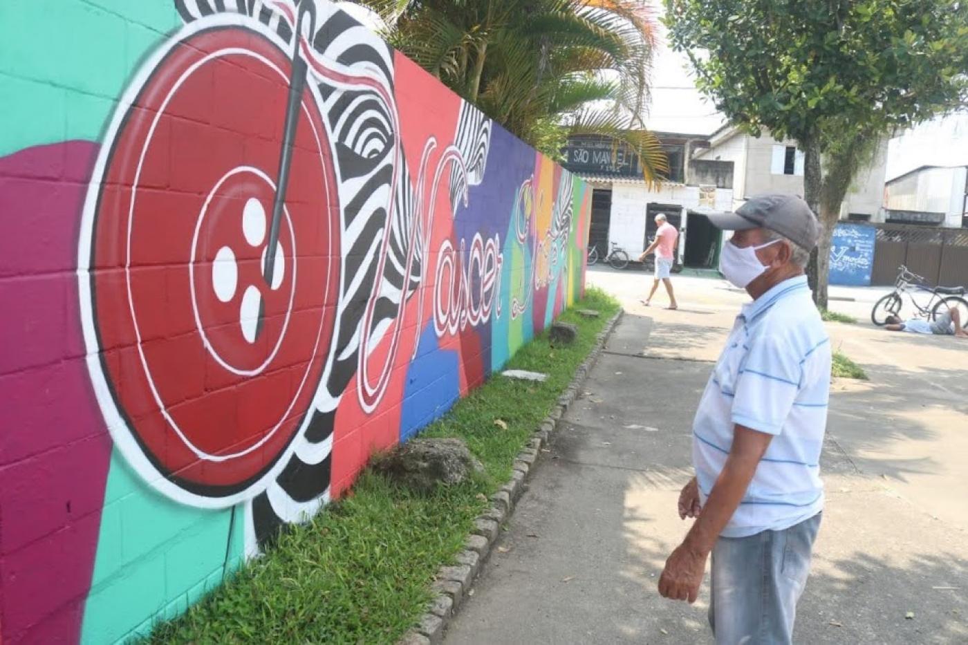 homem em pé na praça usando máscara olha para pintura em muro. . Trecho da calçadaé ajardinado. #paratodosverem