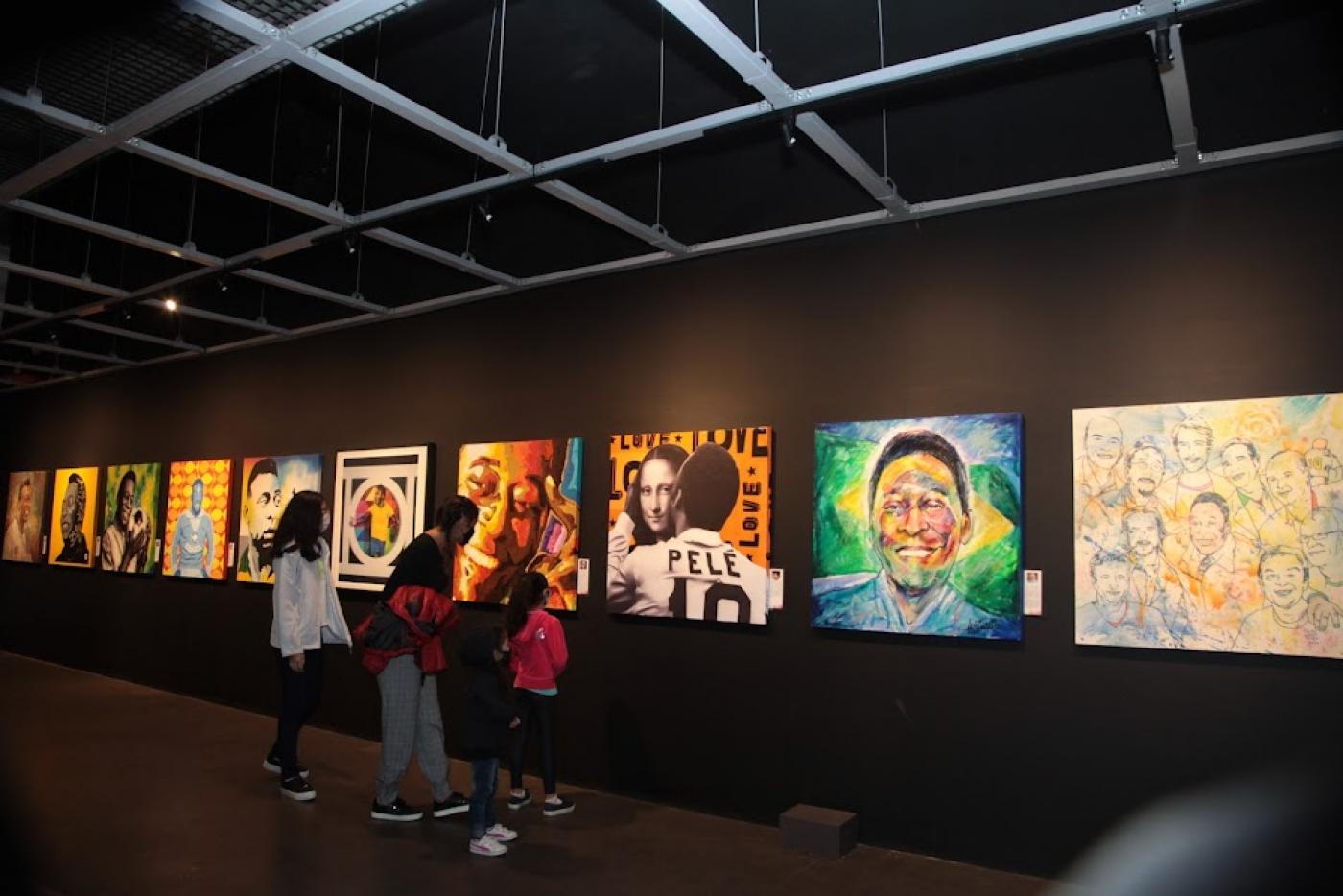 Interior do Museu Pelé com fotos e pinturas do Rei feitas por artistas. Há público observando. #paratodosverem