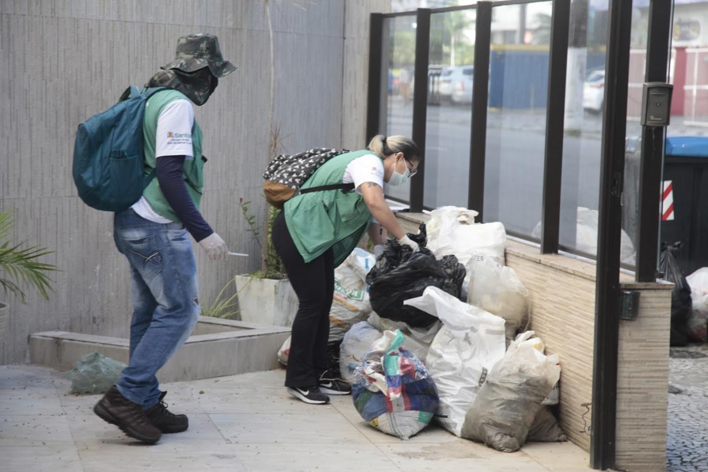 Dois agentes estão em área livre de uma residência. A mulher está vistoriando sacos encostados em um muro. O homem se aproxima dela. #Paratodosverem