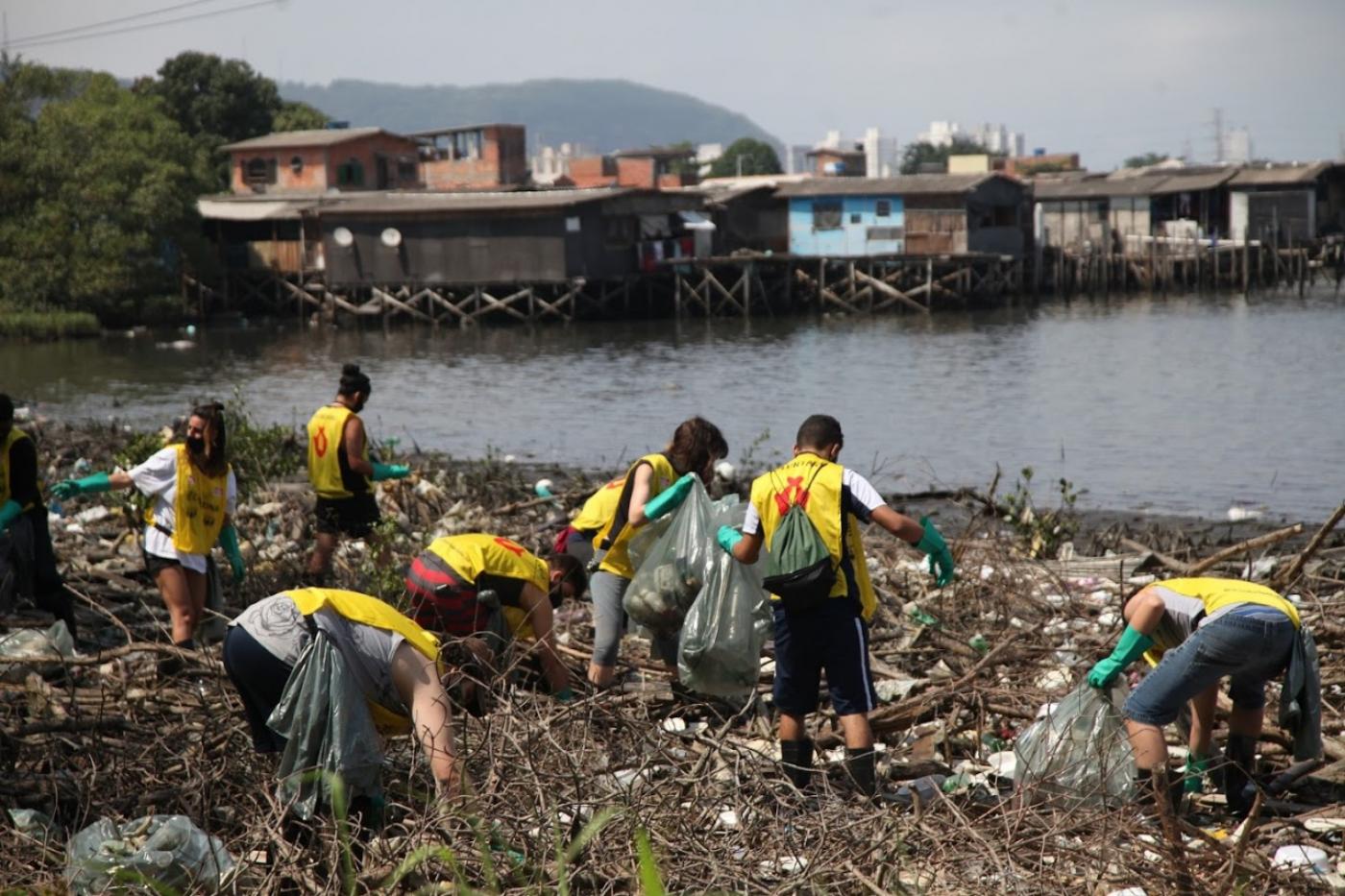 pessoas uniformizadas estão em área de mangue, próximo à água, limpando o terreno. #paratodosverem