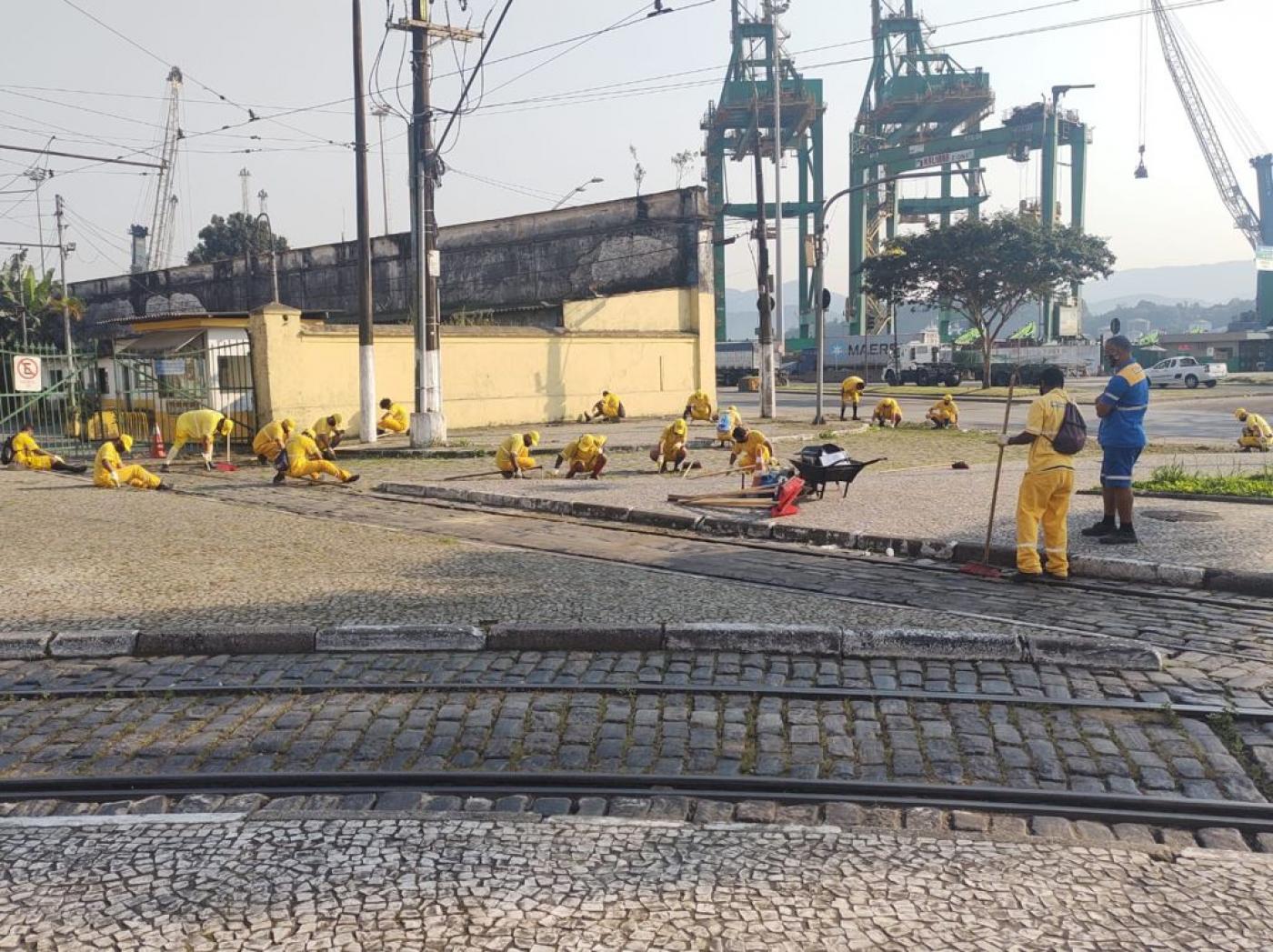 vários homens vestindo uniformes amarelos fazem capinação em calçada. Ao fundo, guindastes no porto. #paratodosverem