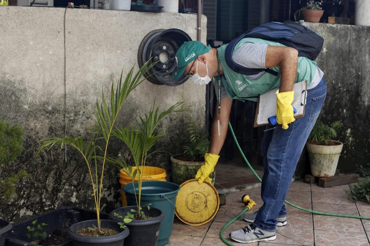 agente uniformizado se curva para pegar prato de planta junto a um jardim. Ele usa luvas nas duas mãos. #paratodosverem