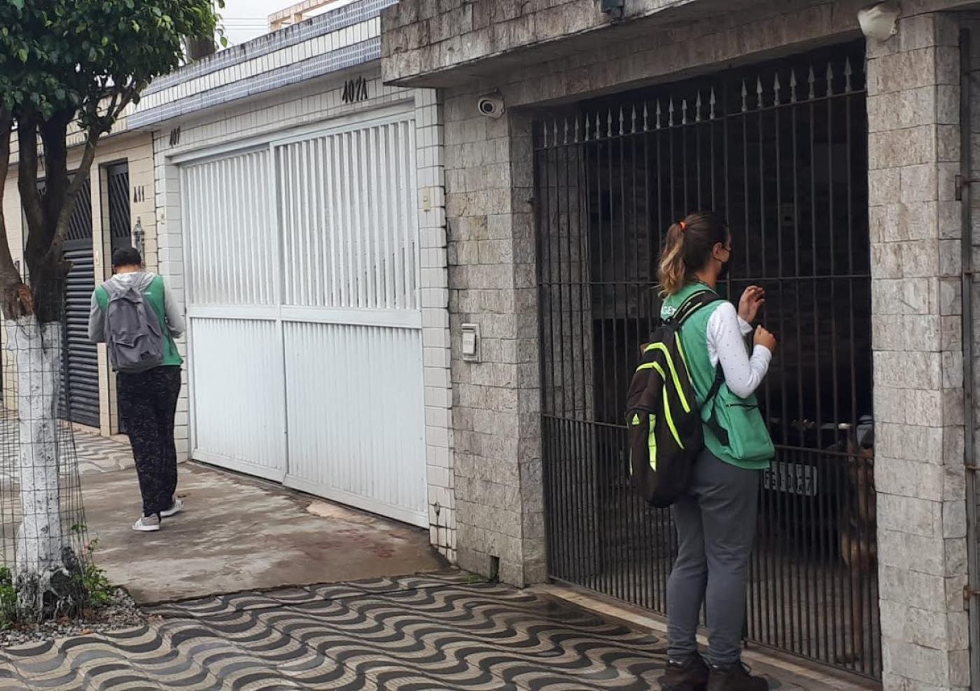 Agente de combate à dengue está à frente do portão de uma casa. Outro agente caminha pela rua. #Paratodosverem