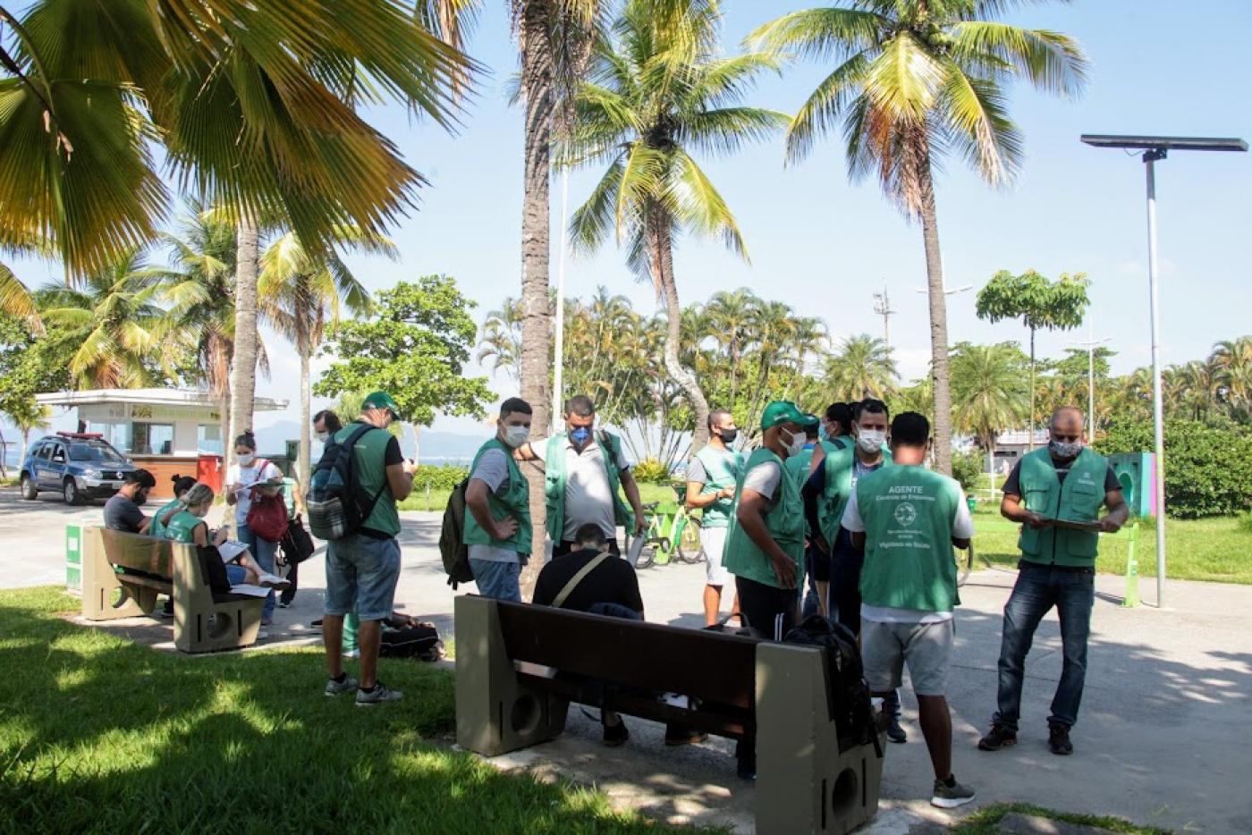 agentes uniformizados estão concentrados no jardim da orla. Em primeiro plano se vê o gramado. Há palmeiras ao redor. #paratodosverem