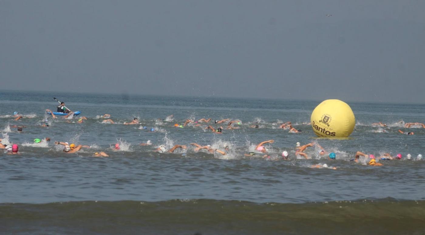 atletas estão nadando no mar. Ao fundo e à direita uma grande boia com o símbolo de santos. #paratodosverem
