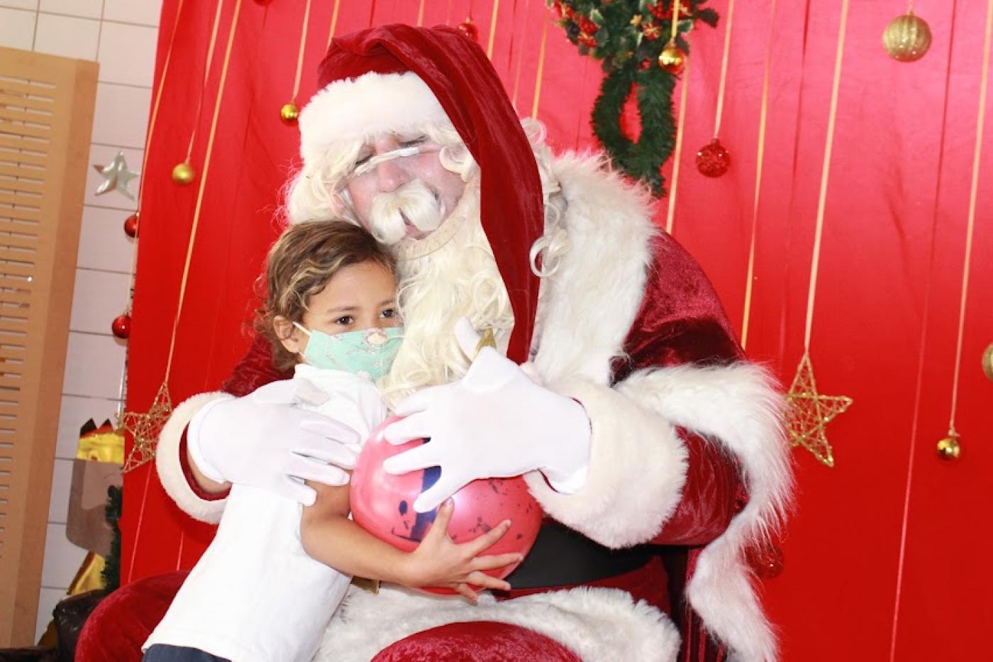 Papai Noel abraça menina. Os dois seguram uma bola colorida. #paratodosverem