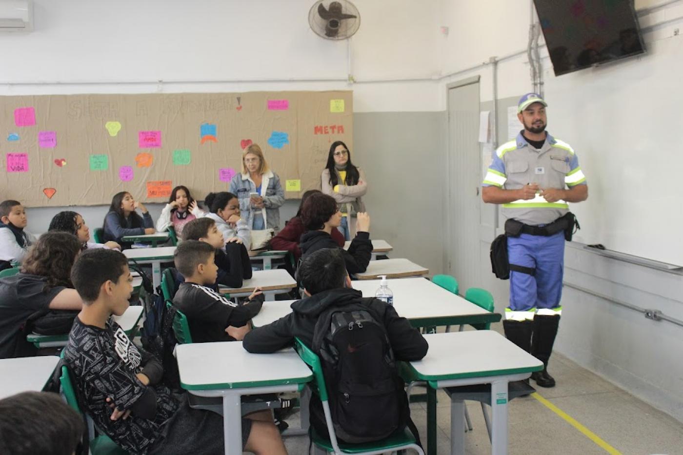 agente de trânsito está à frente de lousa em sala de aula e alunos estão assistindo. #paratodosverem
