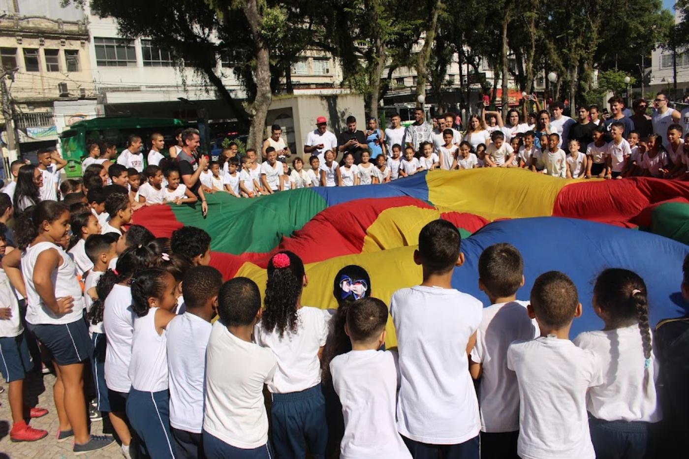 Crianças erguem tecido colorido em movimento conjunto. #paratodosverem
