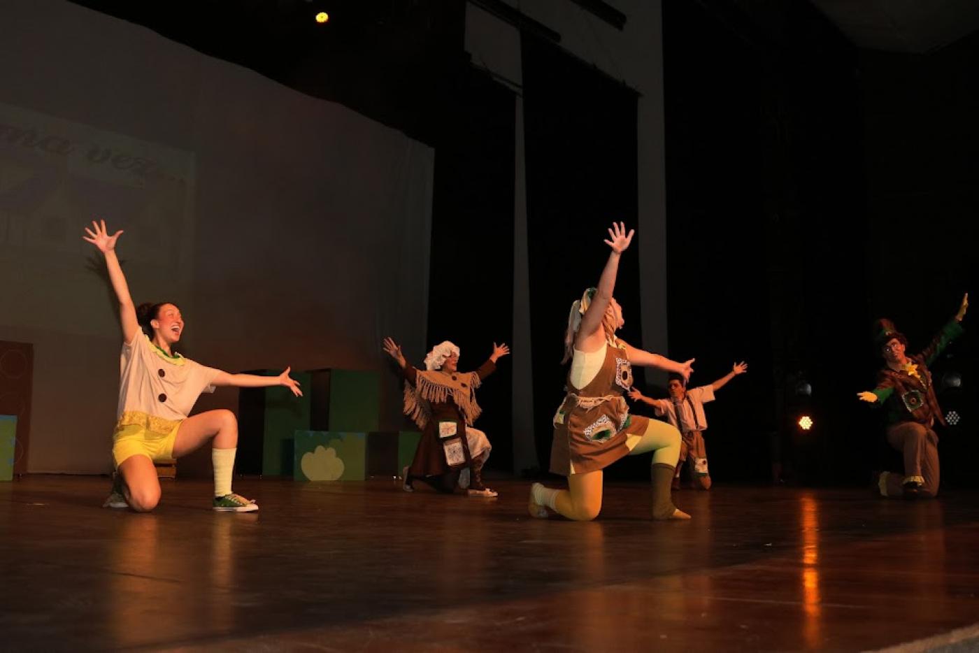 artistas apresentam peça infantil em palco. Eles estão com um dos joelhos apoiados no chão e um dos pés. As mãos estão para o alto. Todos sorriem. #paratodosverem