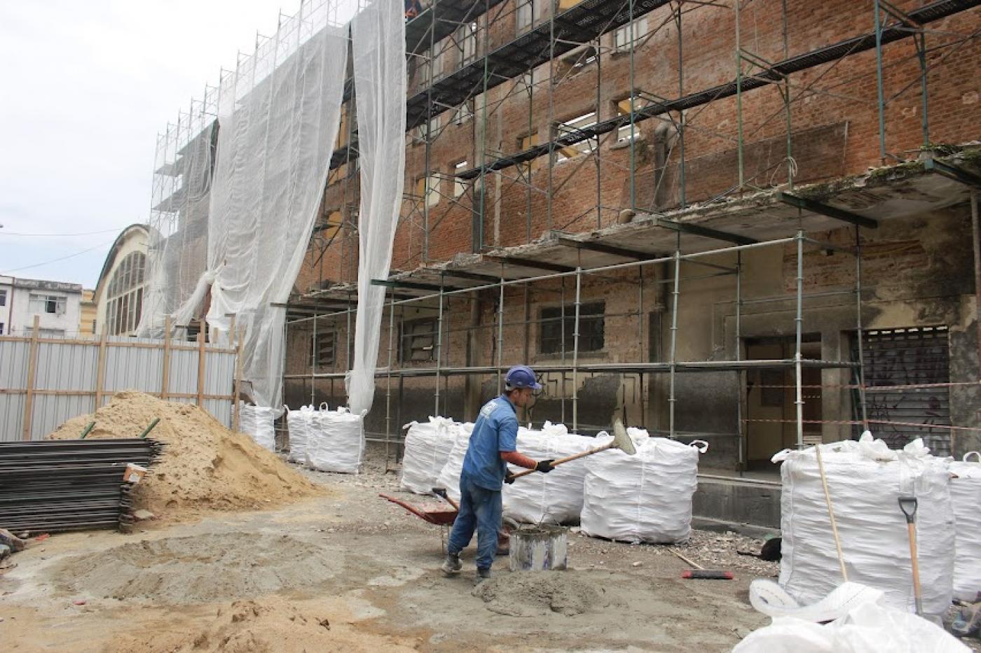 homem está trabalhando em canteiro de obras à frente de uma grande fachada com andaimes. #paratodosverem