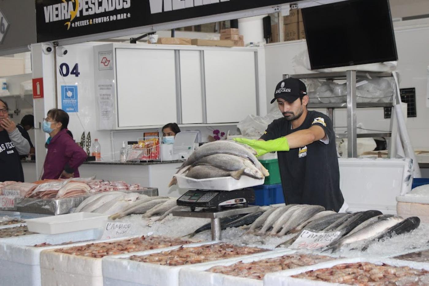 homem pesa em balança vários peixes. À frente dele há várias caixas com pescados. #paratodosverem