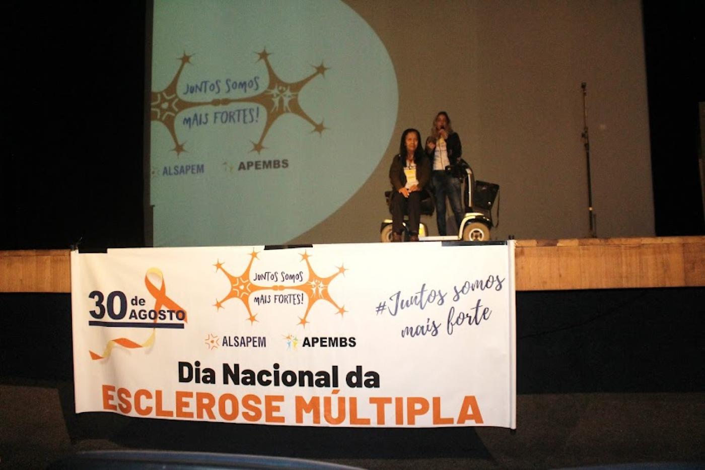 faixa à frente de palco onde se lê Dia Nacional da Esclerose Múltipla. No palco há duas mulheres, uma delas em cadeira de rodas. #paratodosverem