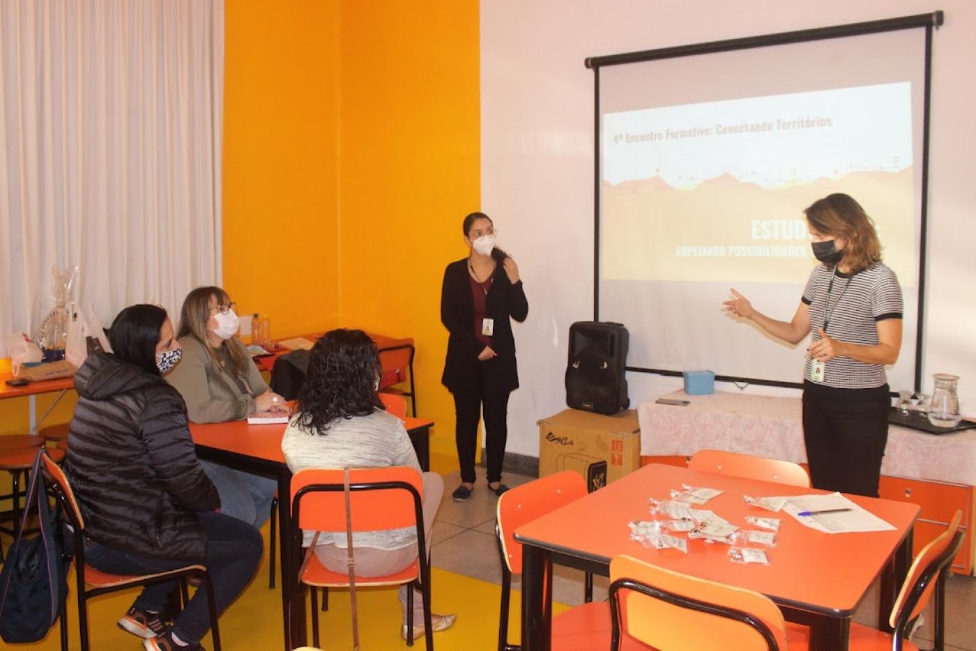 mulher em pé em frente ao telão fala com professoras sentadas #paratodosverem