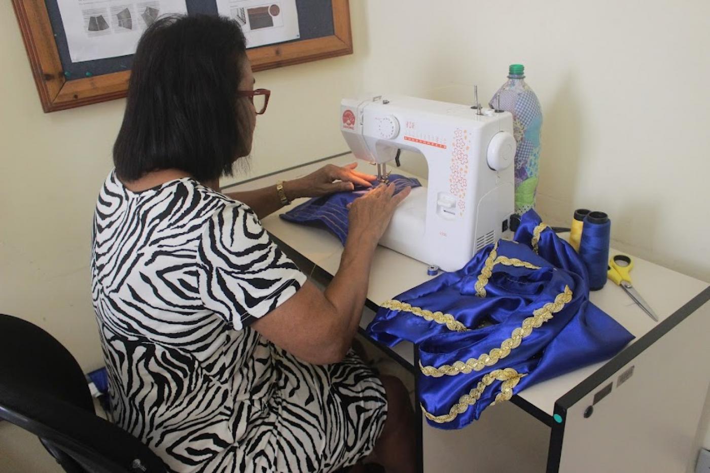 mulher está de costas para a foto confeccionando peça em máquina de costura. Ao lado, um tecido de cetin em azul e dourado. #paratodosverem