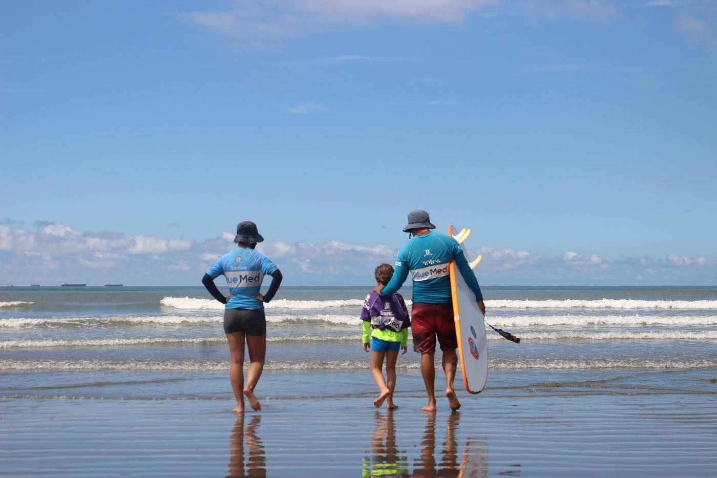criança está entrando no mar com dois adultos. #paratodosverem