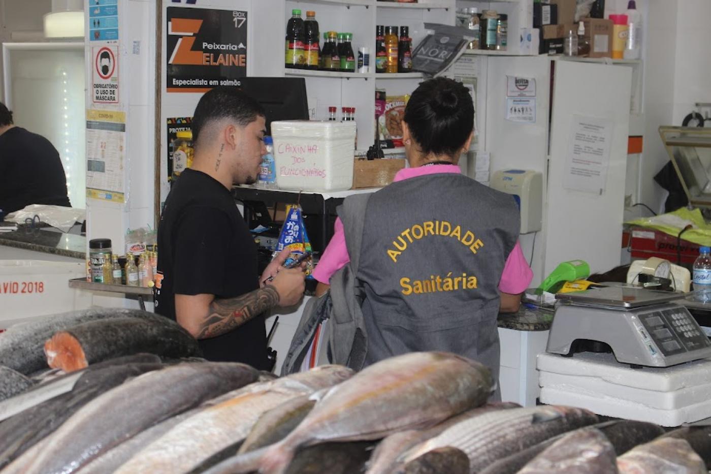 fiscal está de costas conversando com homem em box de peixe. à frente, um balcão cheio de peixes. #paratodosverem 