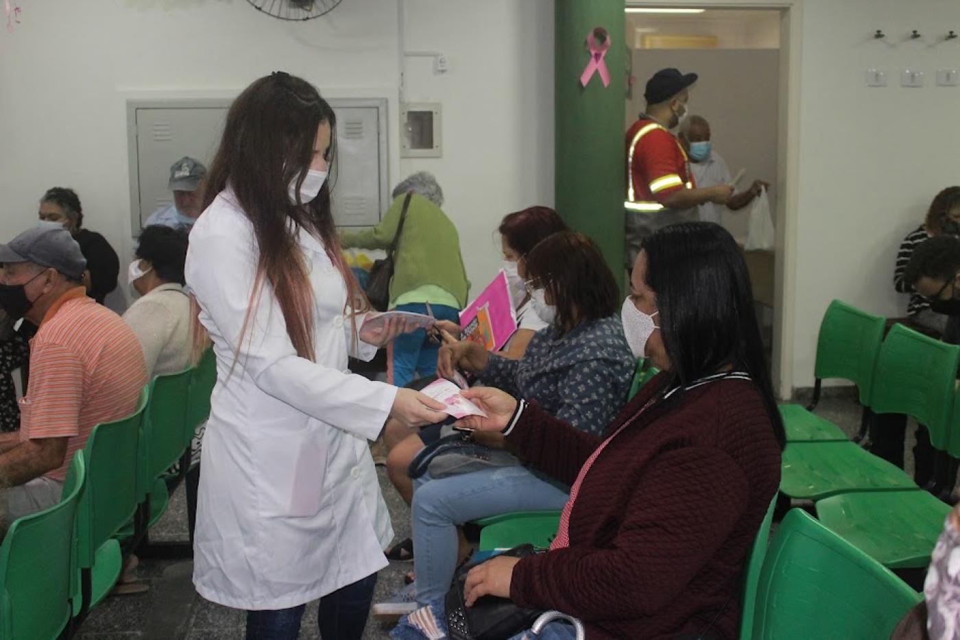 mulher usando avental branco entrega folheto para outra mulher que está sentada em sala de espera. Outras pessoas estão sentadas também. #paratodosverem