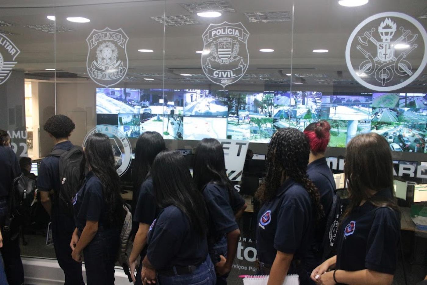 jovens todos de costas para a foto olham para sala de monitoramento do Centro de Controle Operacional. #paratodosverem