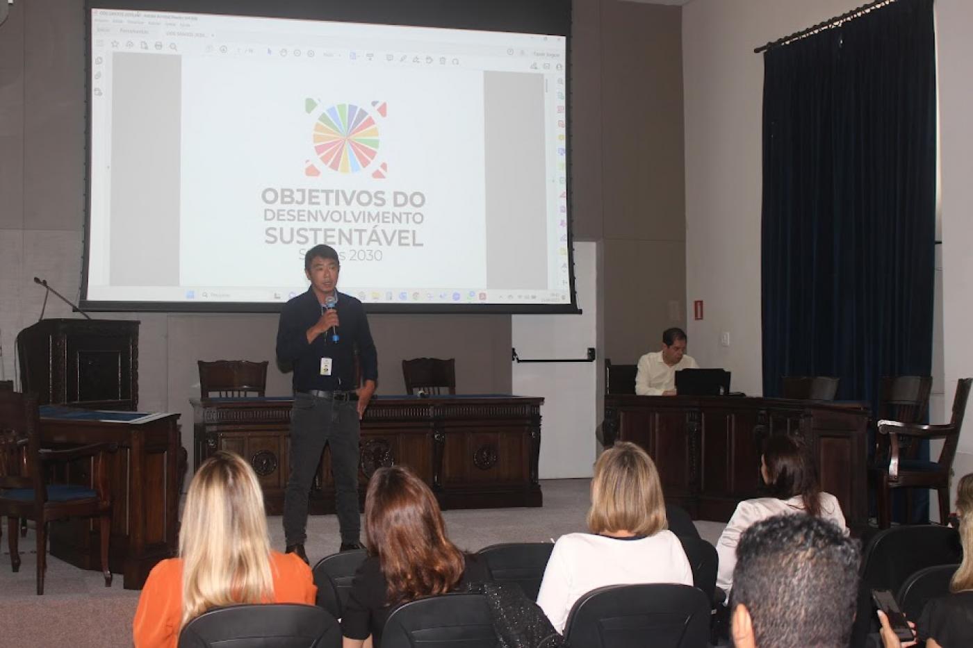 homem está falando diante de telão onde se lê objetivos do desenvolvimento sustentável. #paratodosverem 