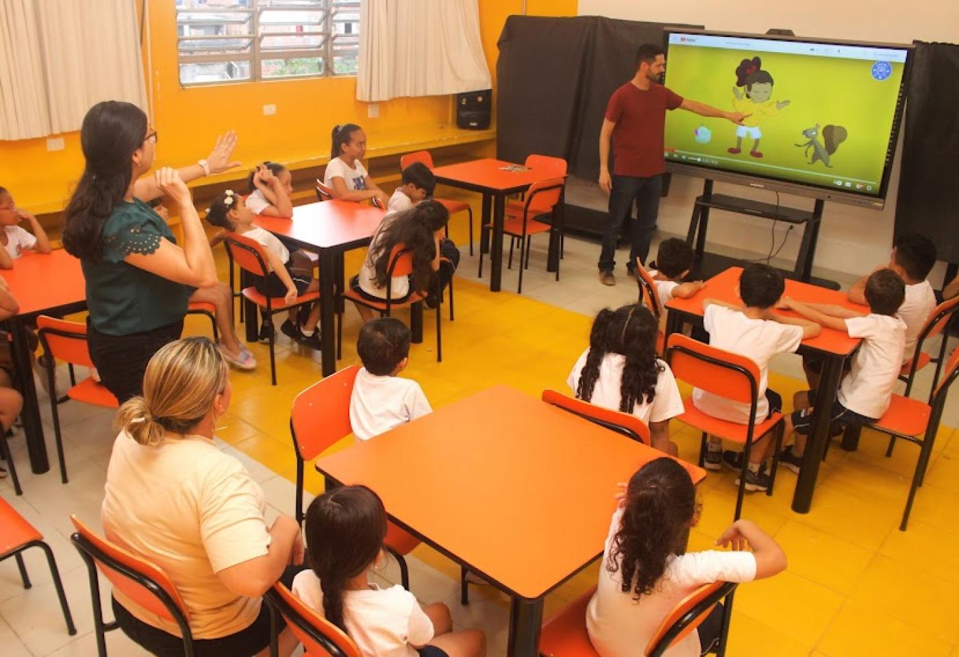professor está diante de tela apontando para figura e crianças estão sentadas prestando atenção. #paratodosverem