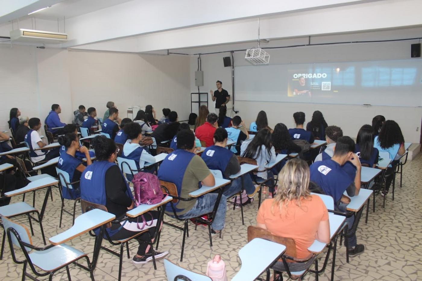 pessoas estão sentadas em sala de aula. todos estão de costas para a foto. Ao fundo, um homem fala ao grupo à frente de um telão. #paratodosverem